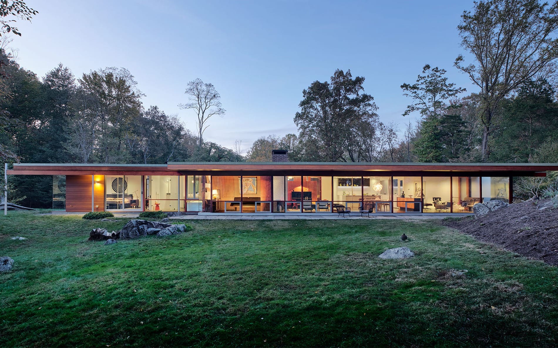 Modern glass house in lush forest setting at dusk, showcasing open interiors and expansive glass walls.