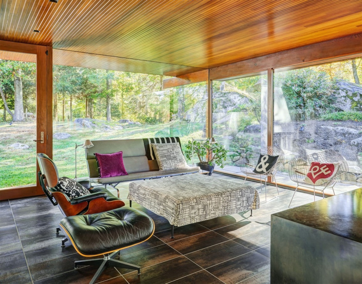 Modern living room with large windows, stylish furniture, and a view of a lush forest outside. Warm lighting and decor.