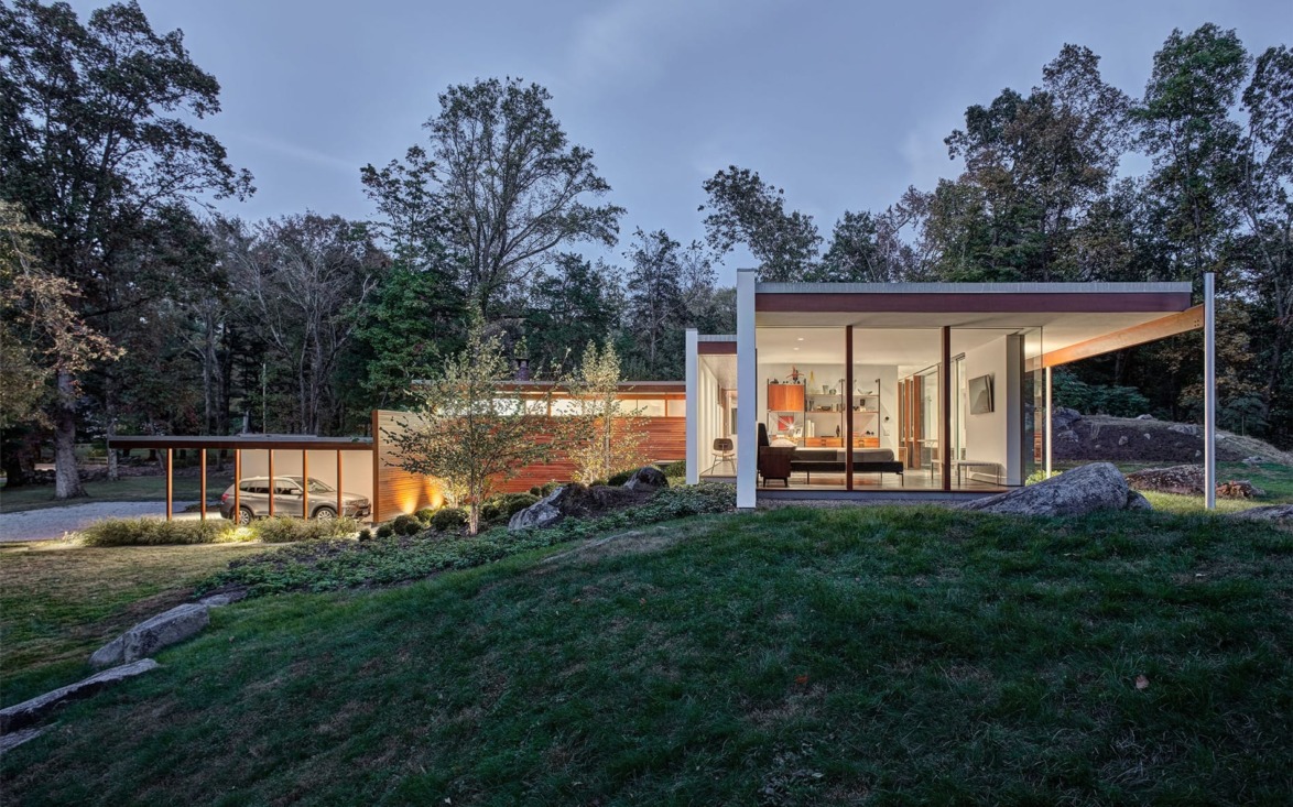 Modern glass-walled house surrounded by trees, illuminated at dusk, with a spacious lawn in the foreground.