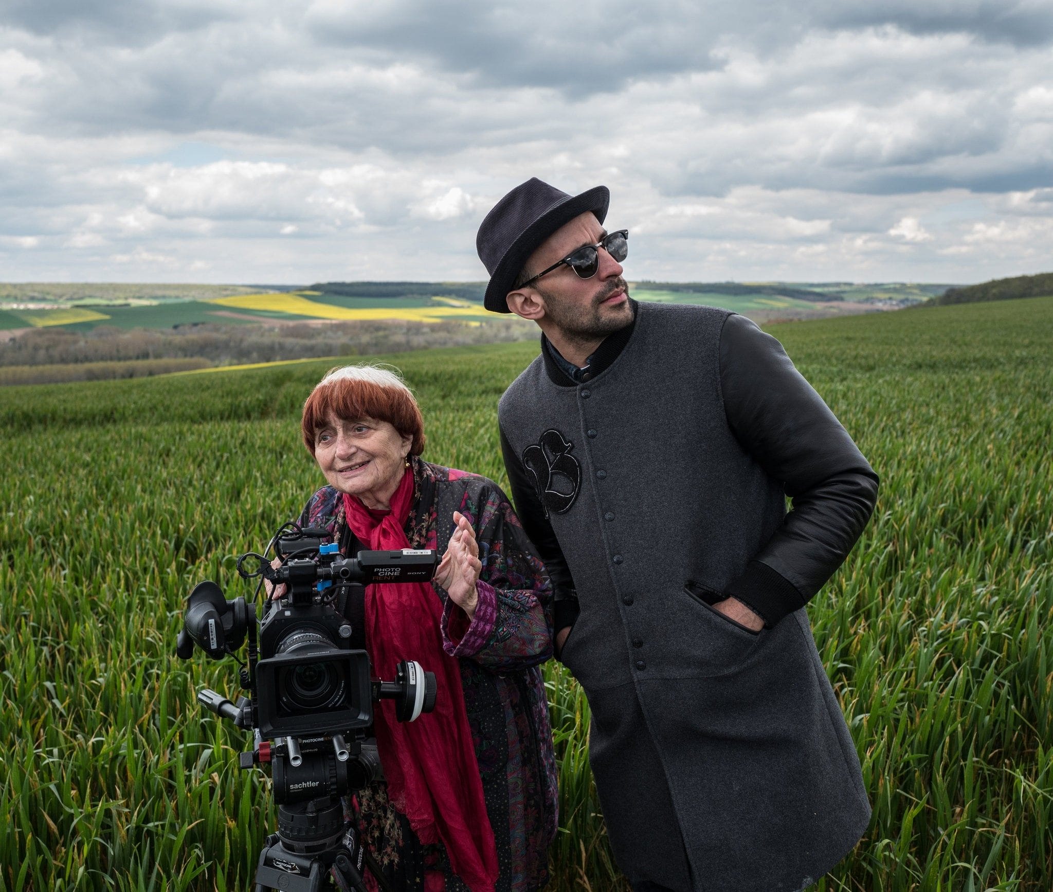 Two individuals stand in a field beside a film camera, with rolling hills and a cloudy sky in the background.