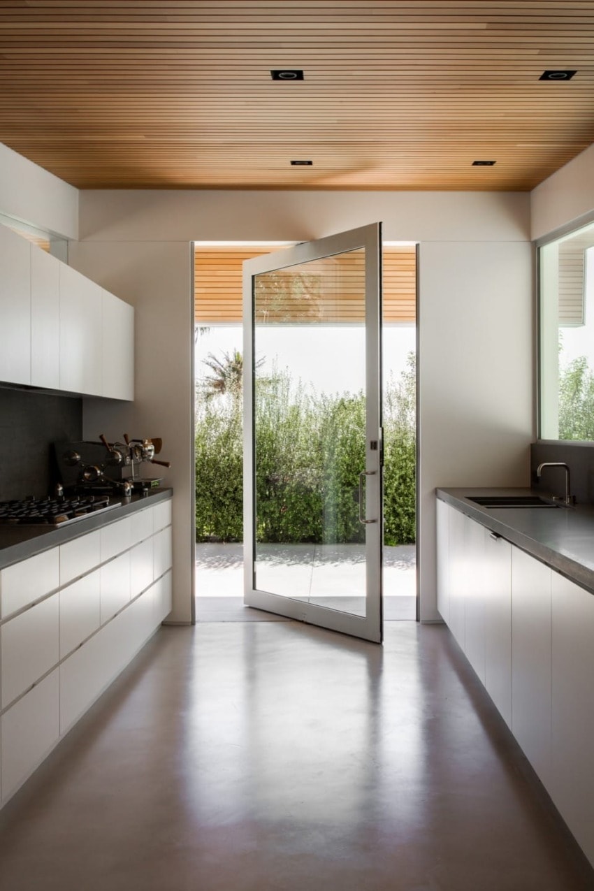Modern kitchen with sleek white cabinetry, large glass door open to a garden view, and wooden ceiling accents.