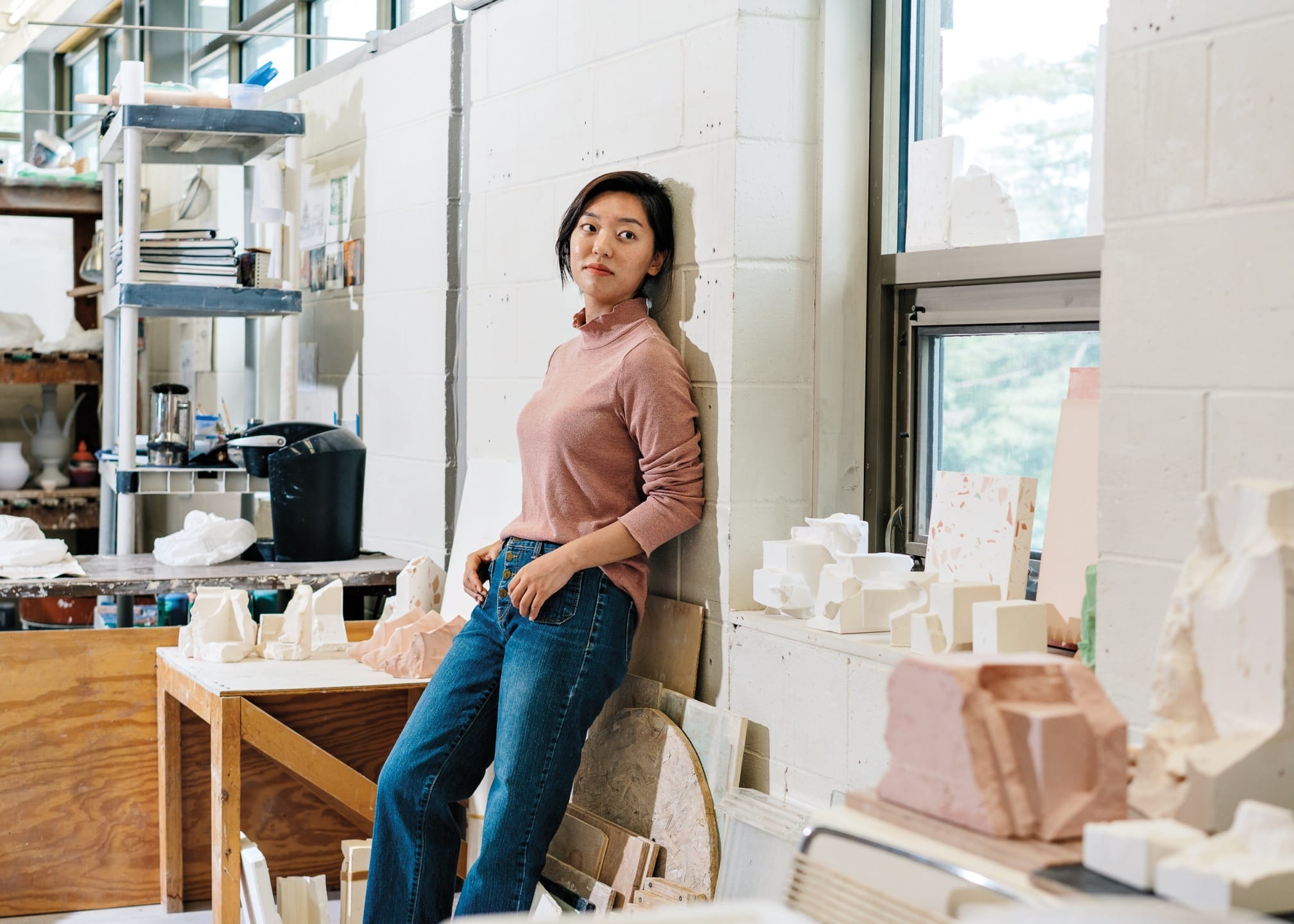 Person leaning against wall in art studio surrounded by sculptures and art supplies.