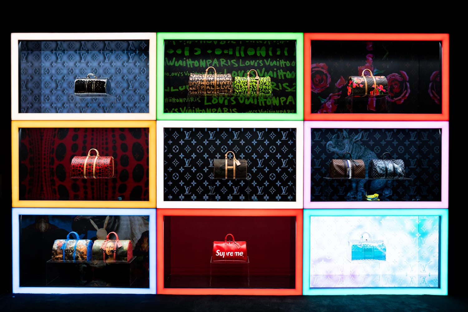 Colorful display of designer handbags in neon-lit boxes on a black wall background.