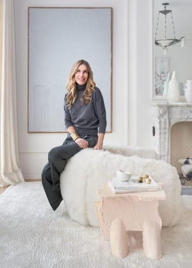 Person sitting on a cozy chair in a stylish, light-filled living room with modern decor and a marble fireplace.