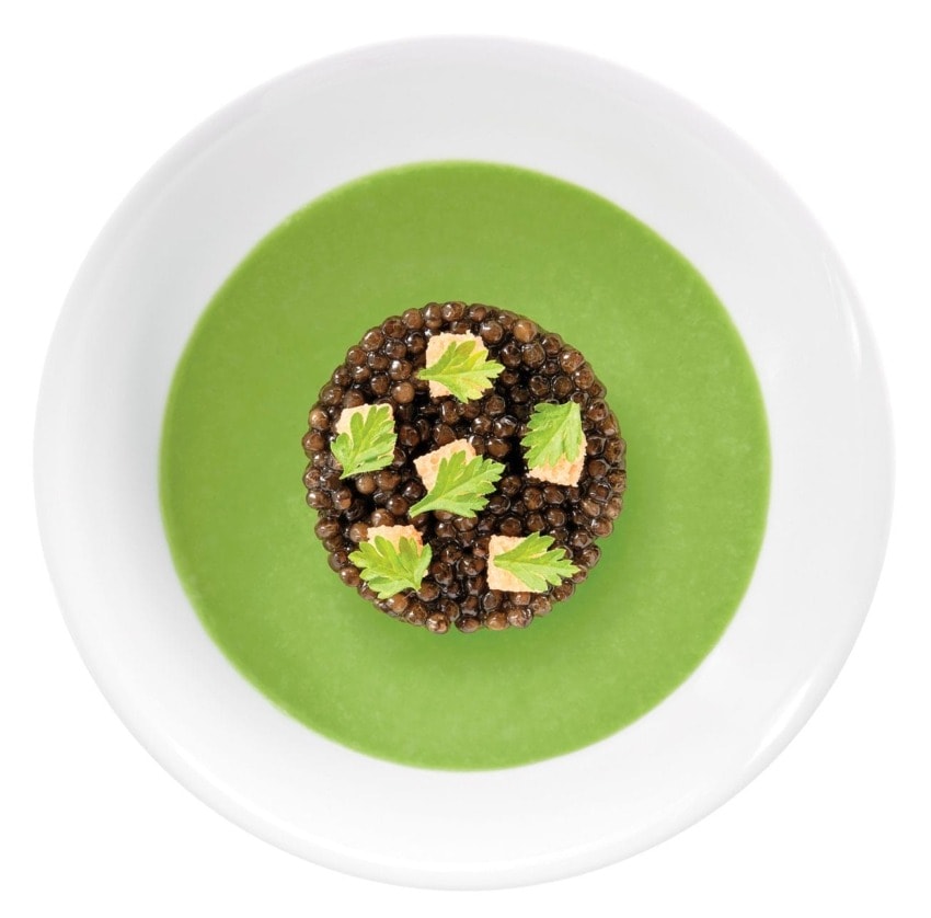 Top view of a elegant dish featuring green soup with a central garnish of lentils and herbs in a white bowl.