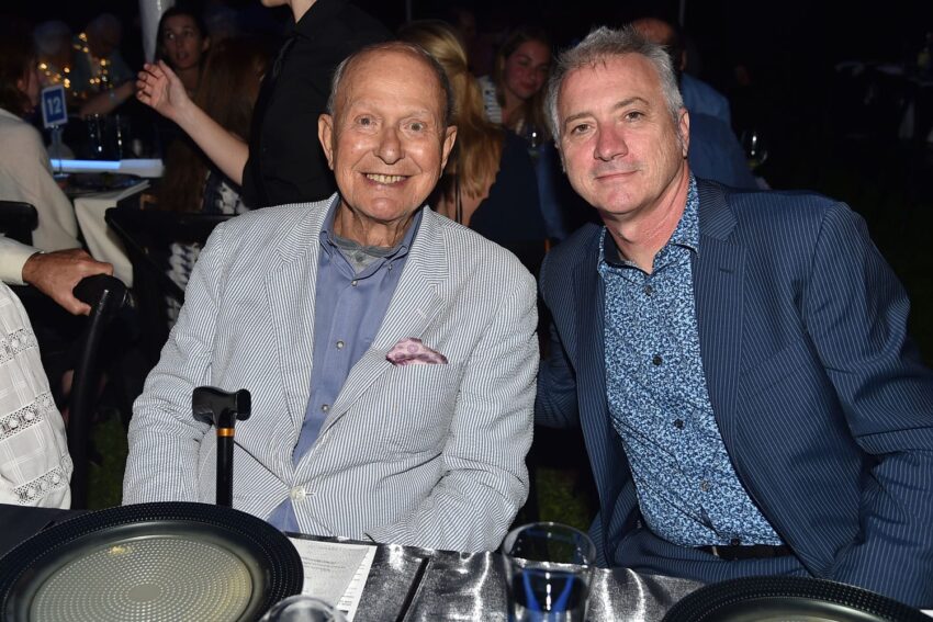 Two men seated at a dining event, smiling at the camera, with plates and cutlery on the table in front of them.