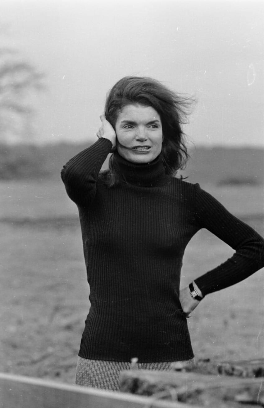 Woman in a turtleneck sweater outdoors with windblown hair, hand touching head, standing in a field.