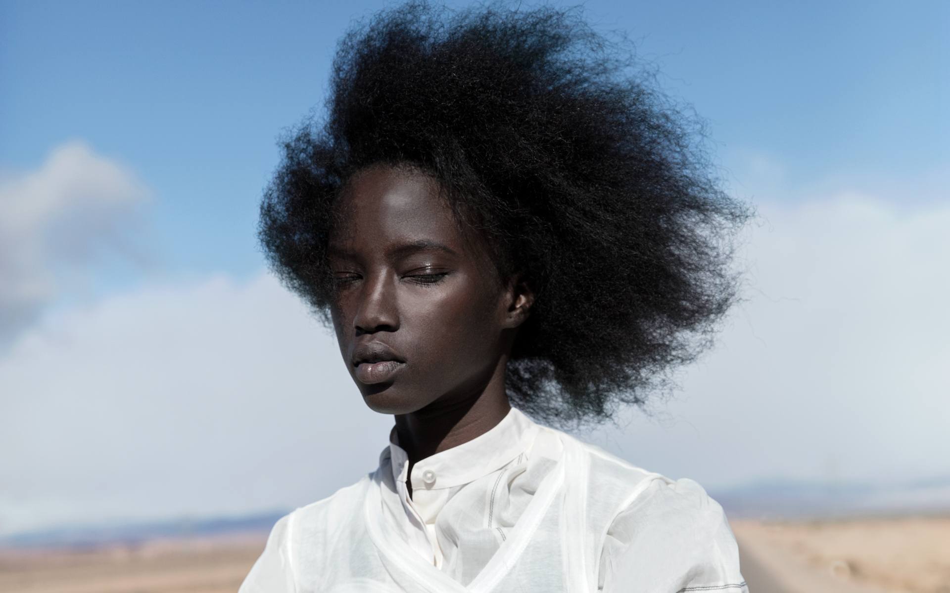 Woman in a white blouse standing in a desert landscape with a clear blue sky and wild, voluminous hair.