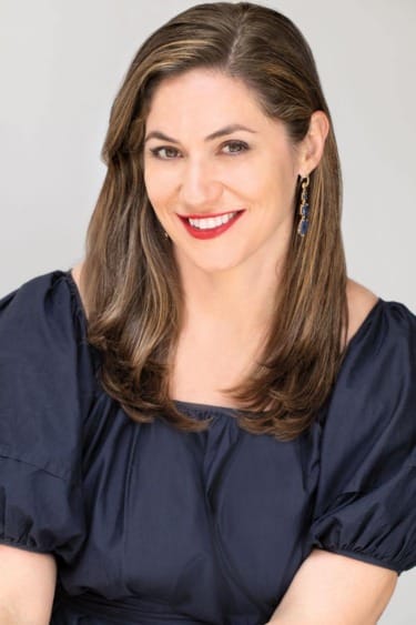 Person smiling, wearing a dark blouse and earrings, with long brown hair against a light background.