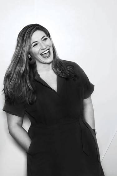 Woman in a dark dress laughing with hands in pockets against a white background.