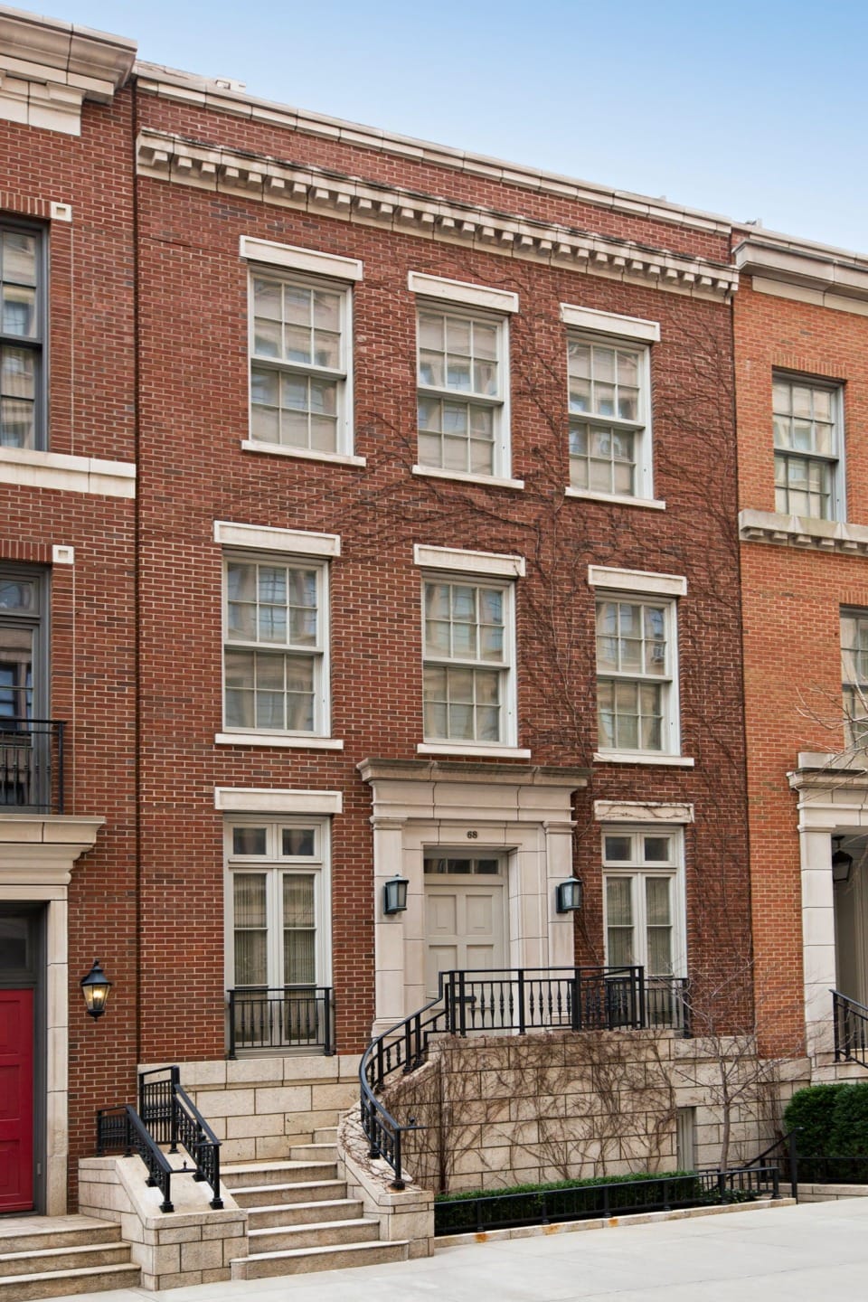 Red brick townhouse with white trim, black railings, and a central door, surrounded by stone steps and neighboring buildings.