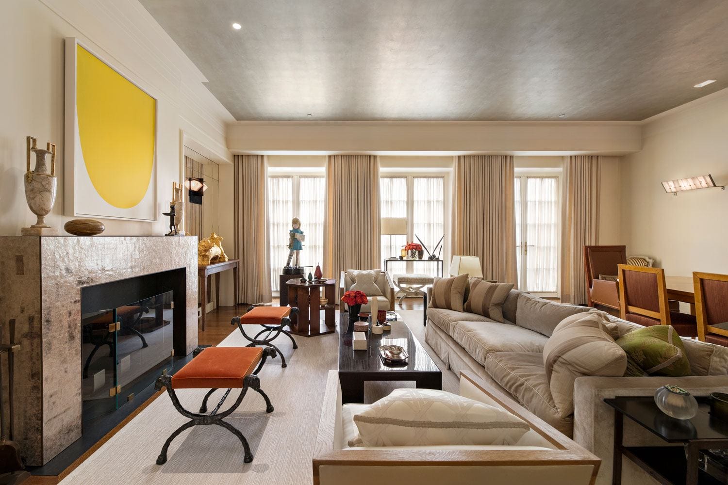Modern living room with beige sofas, a yellow art piece above the fireplace, and large windows with curtains.