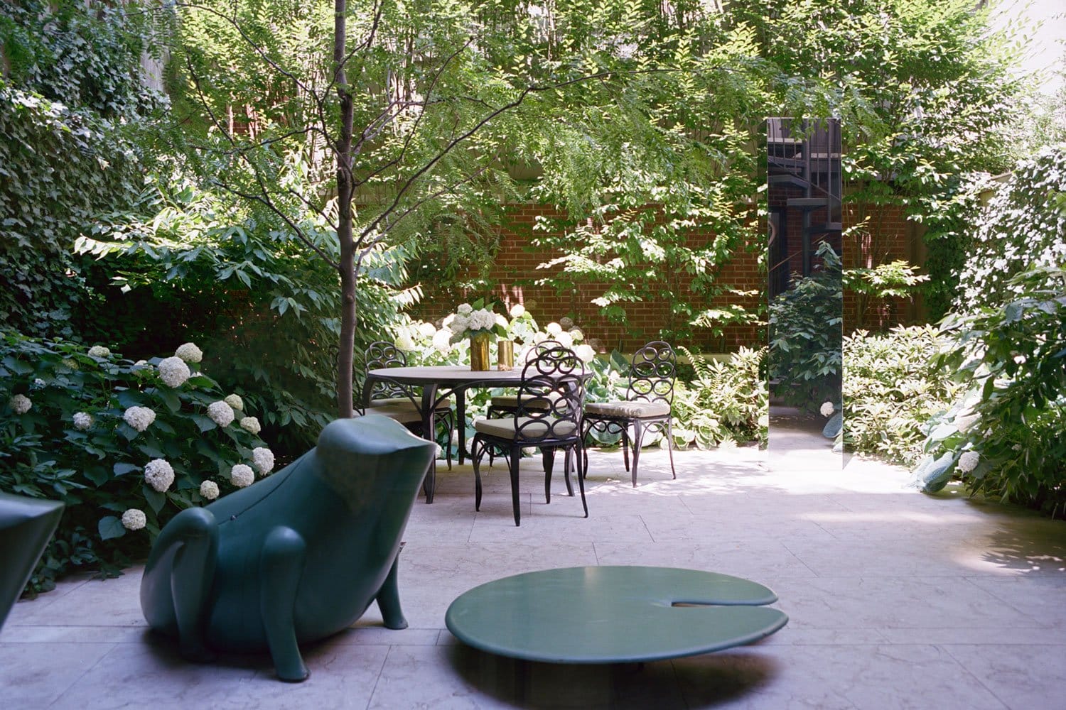 Outdoor garden with modern green furniture and lush greenery, featuring a table, chairs, and decorative sculptures.