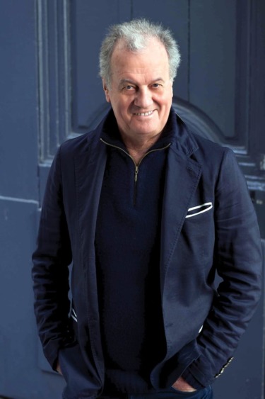 Smiling man in a dark jacket standing in front of a blue door background.