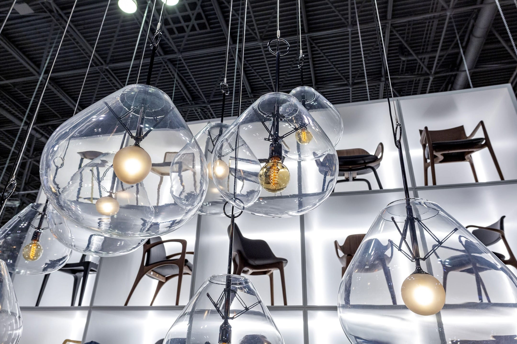 Modern hanging light fixtures with clear glass and visible bulbs on display in a showroom with chairs in the background.