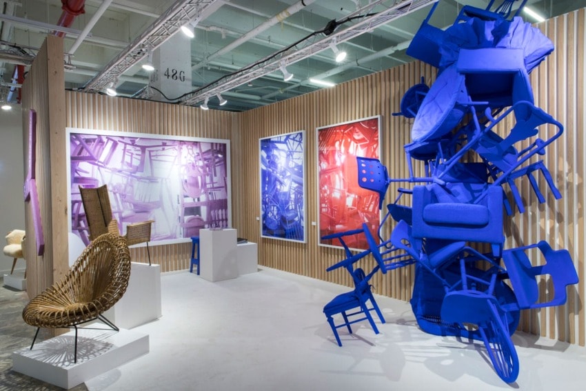 Art exhibit featuring a vibrant installation of blue chairs, rattan furniture, and large colorful framed prints on the wall.