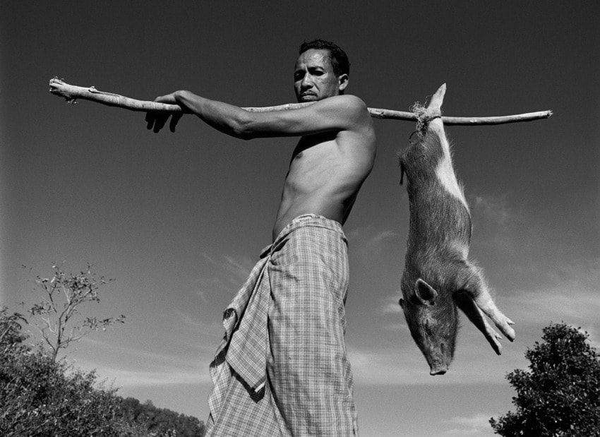 Man carrying a wild boar on a stick over his shoulders, standing shirtless in a rural outdoor setting, black and white photo.