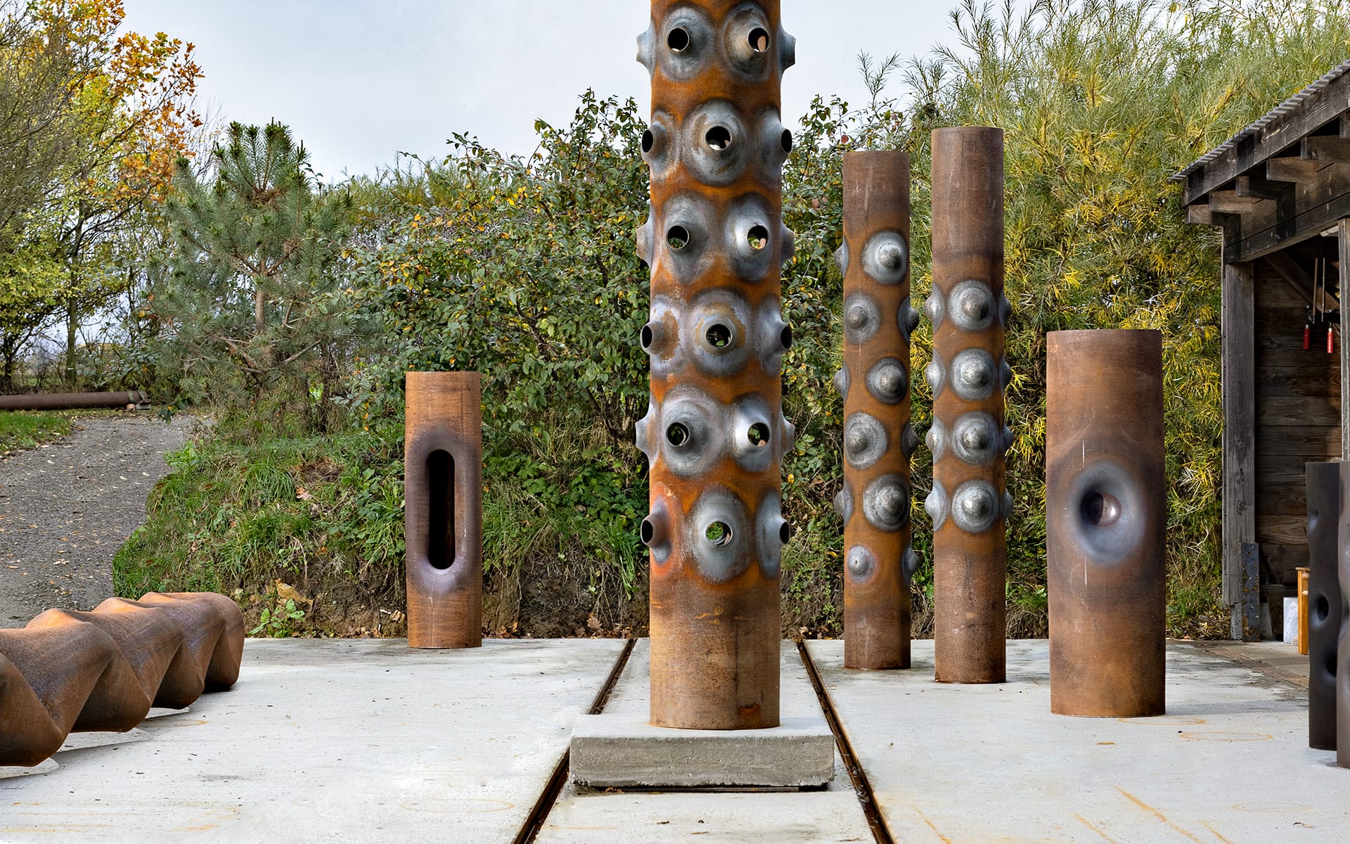 Outdoor art installation with tall, rust-colored metal columns featuring unique textures and shapes, surrounded by greenery.