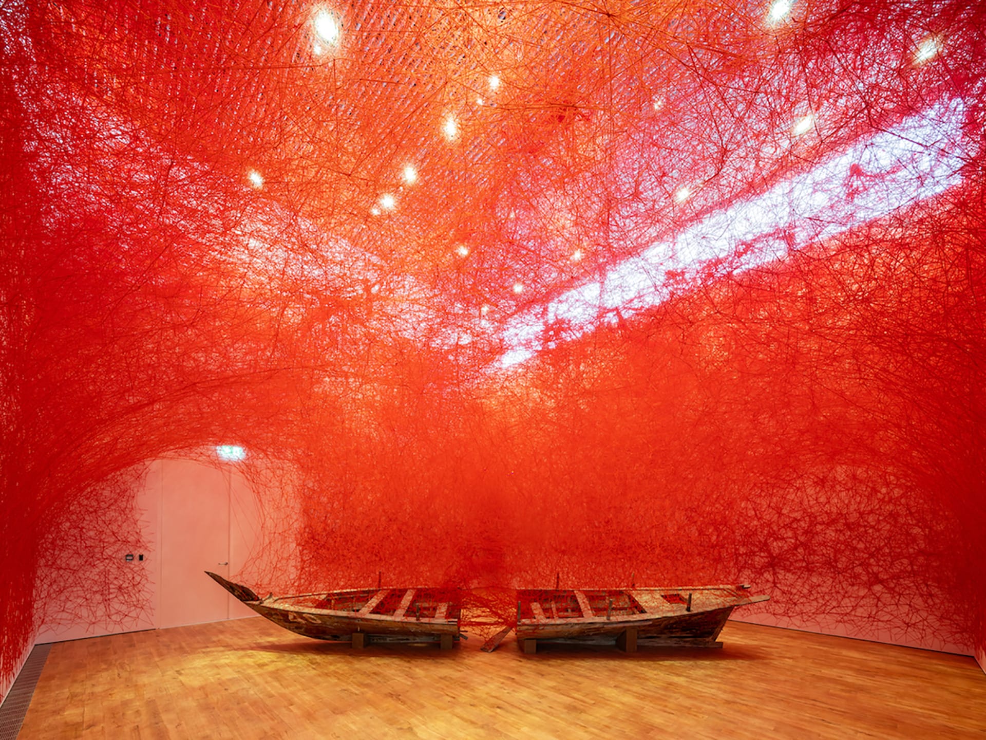Art installation with two wooden boats surrounded by intricate red threads creating a vibrant, immersive environment in a gallery.