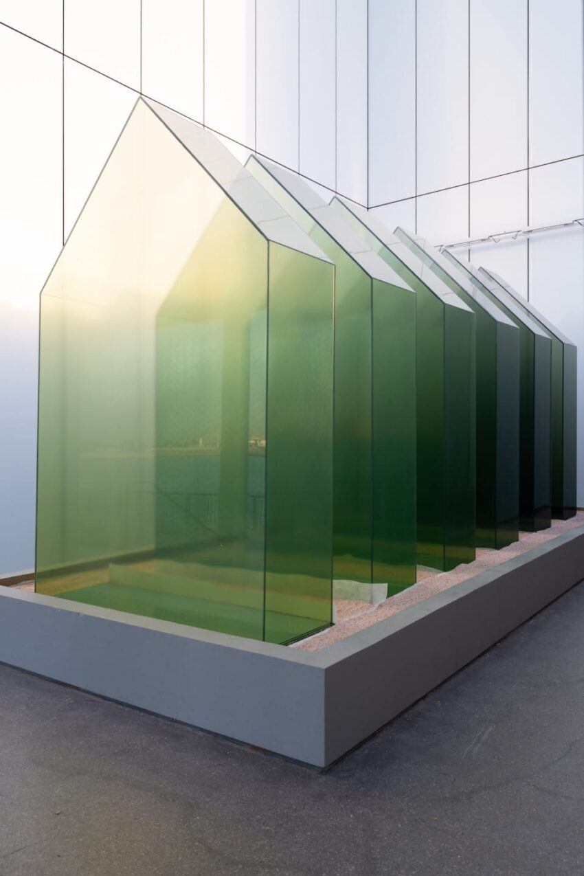 Art installation of several translucent green glass structures resembling small houses on a concrete platform indoors.