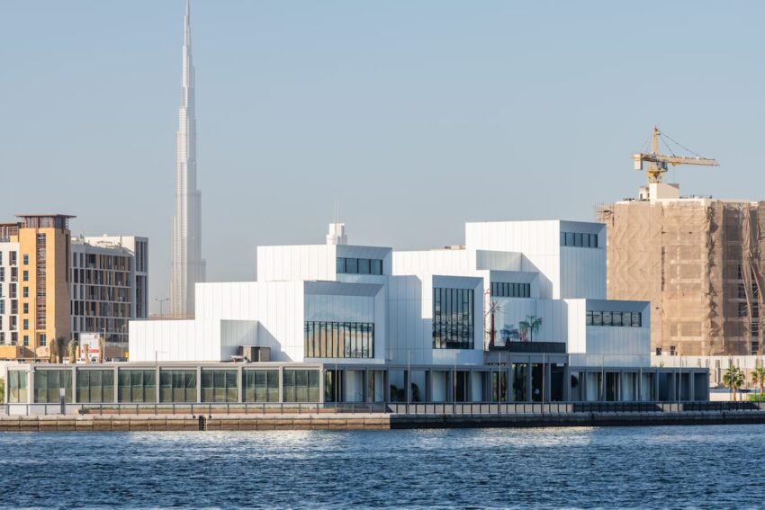 Modern white building on waterfront with cityscape and crane in the background on a clear day.