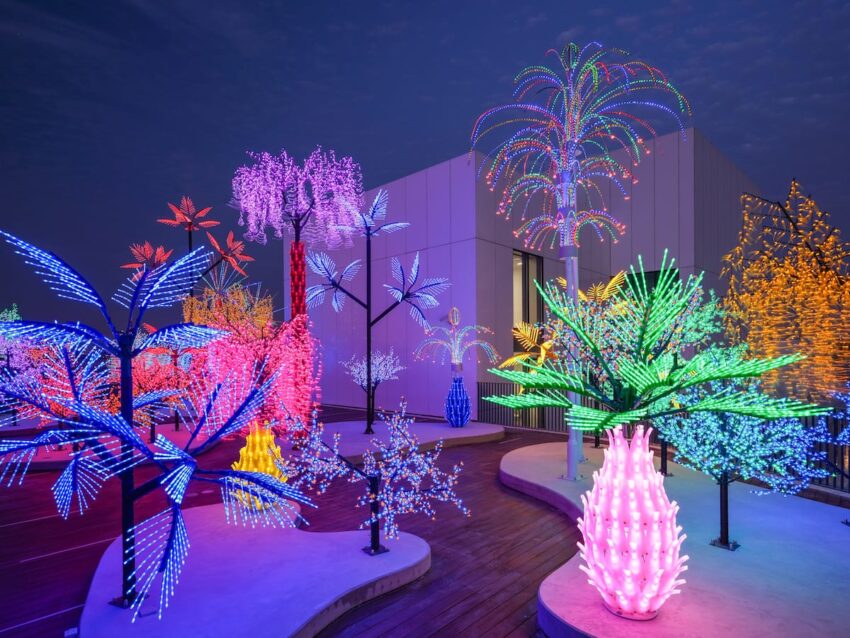 Illuminated display of vibrantly colored LED trees with various shapes and sizes against a modern building at night.