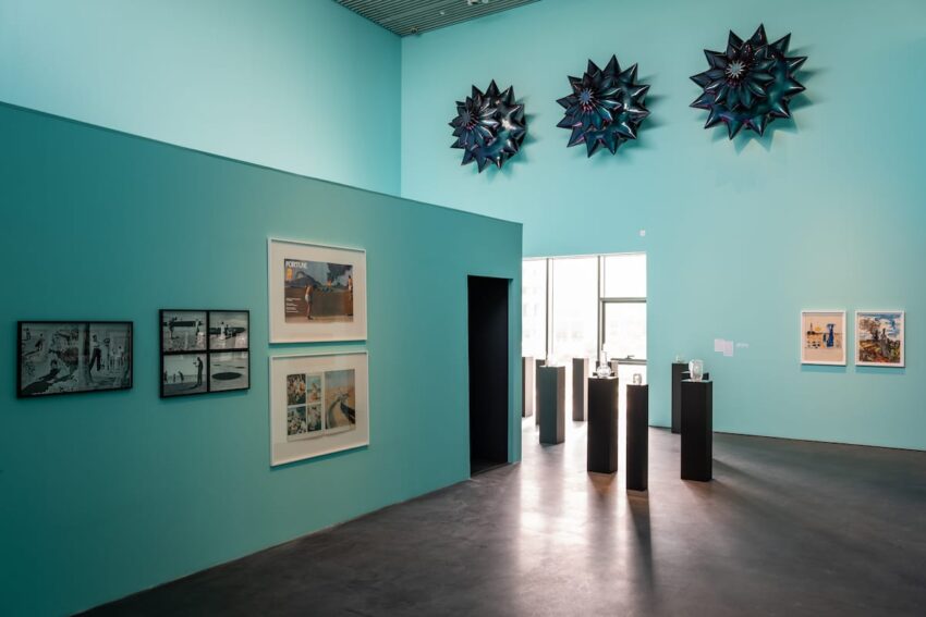 Art gallery interior with teal walls displaying framed photographs, sculptures on pedestals, and star-shaped wall installations.