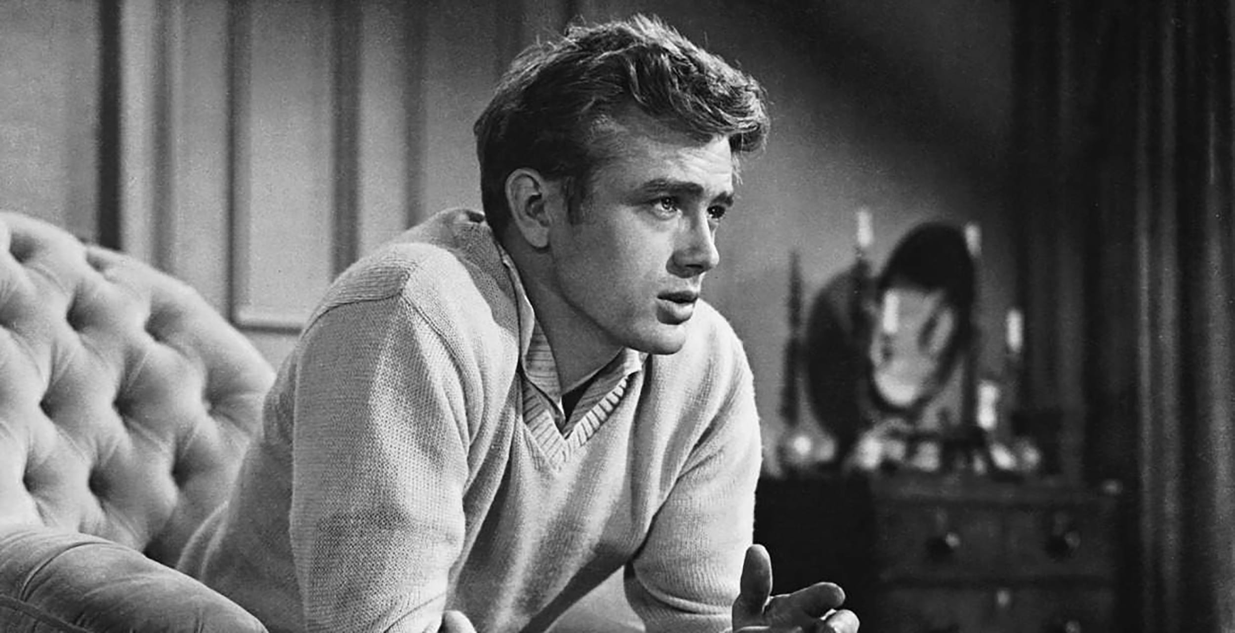 Black and white photo of a contemplative man sitting in a living room, wearing a sweater, with hands clasped together.