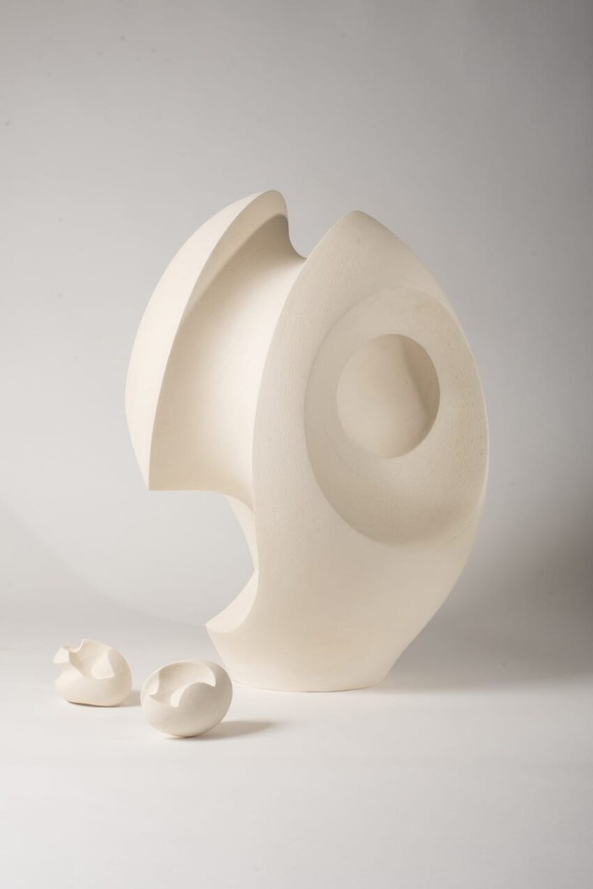 Abstract white sculpture with smooth curves and hollow center, accompanied by two small similar shapes on a plain background.