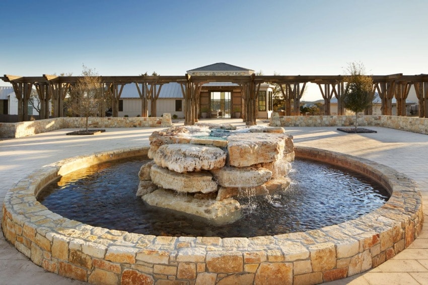 Stone fountain in a circular courtyard with a wooden pergola and building in the background under a clear blue sky.