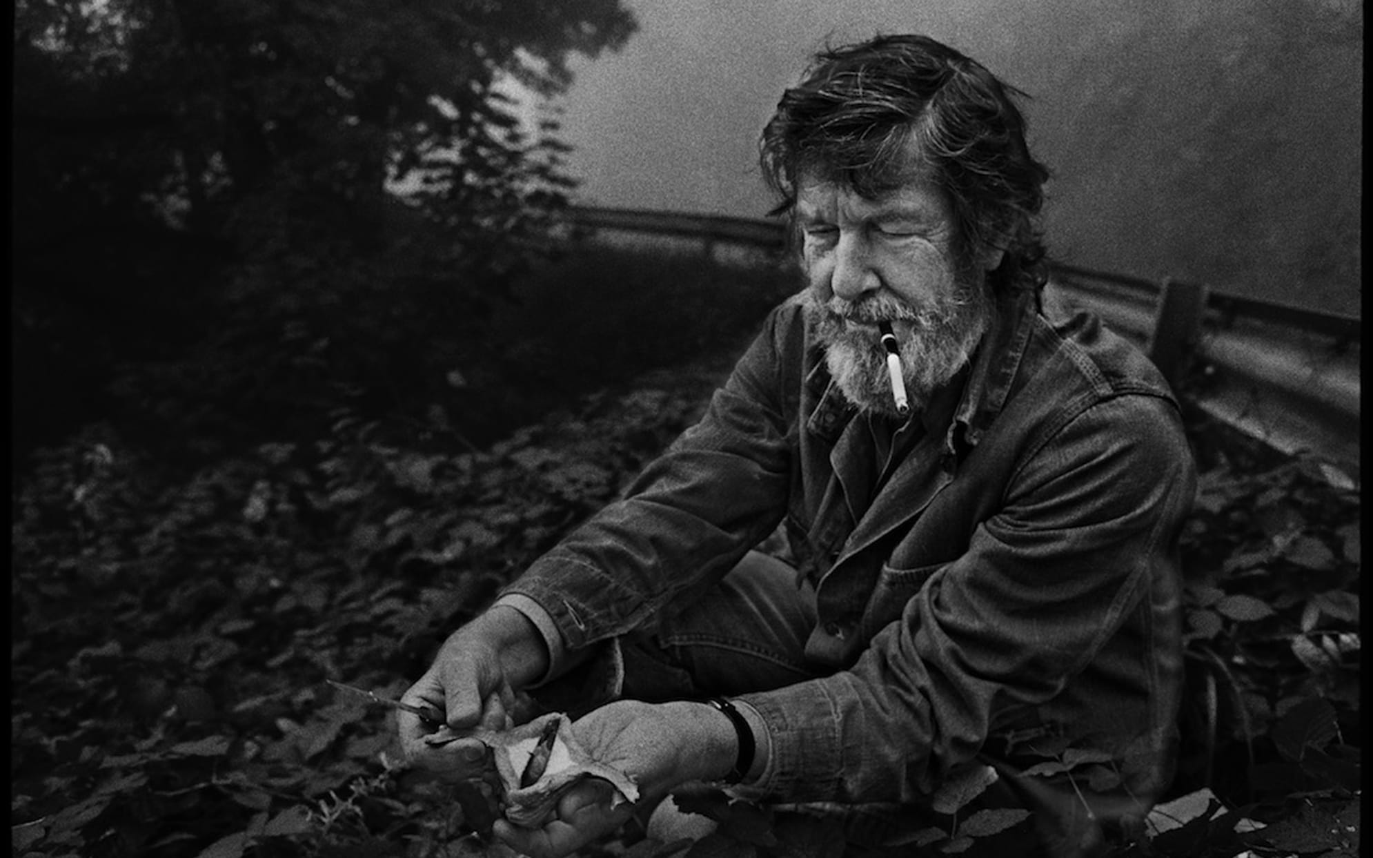 Bearded man sitting outdoors, smoking a pipe, and cleaning mushrooms with a serene expression amidst foliage.