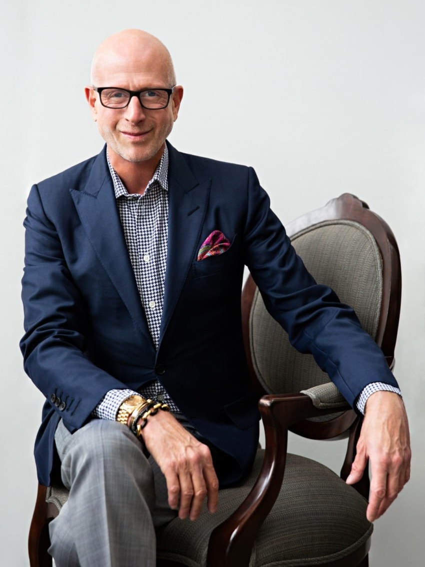 Bald man in glasses and navy blazer sitting on a chair, wearing checkered shirt and colorful pocket square, smiling at camera.