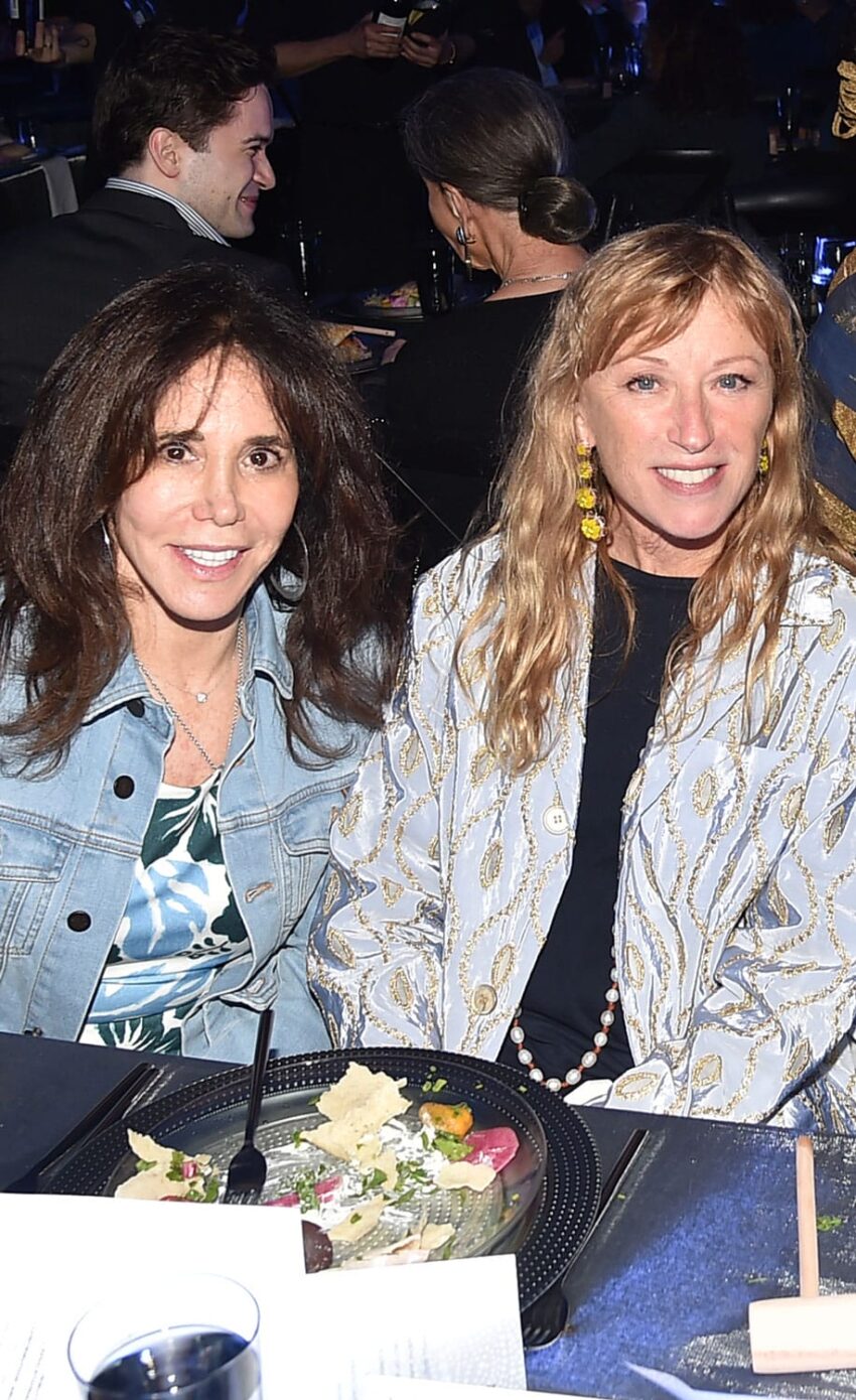 Two women smiling and seated at a dining table with a partially eaten salad in front of them at an indoor event.
