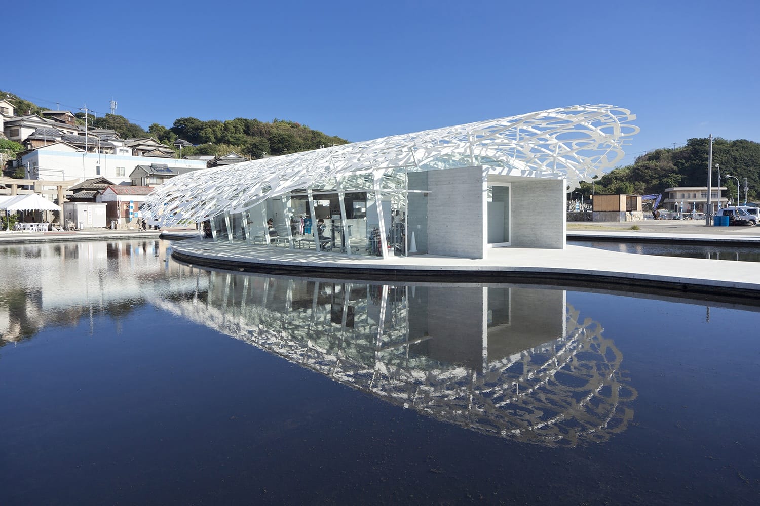 Modern glass and steel pavilion with intricate design on a clear day, reflecting in calm water, surrounded by trees and houses.