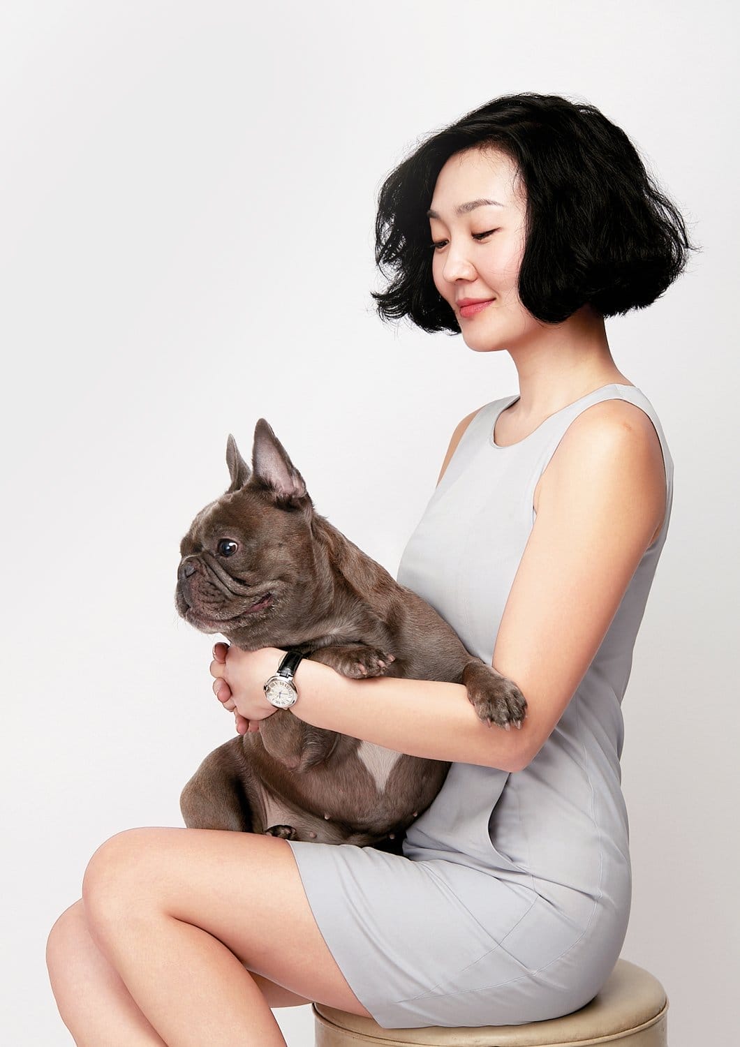 Woman in a gray dress sitting on a stool, holding a French bulldog, against a white background.
