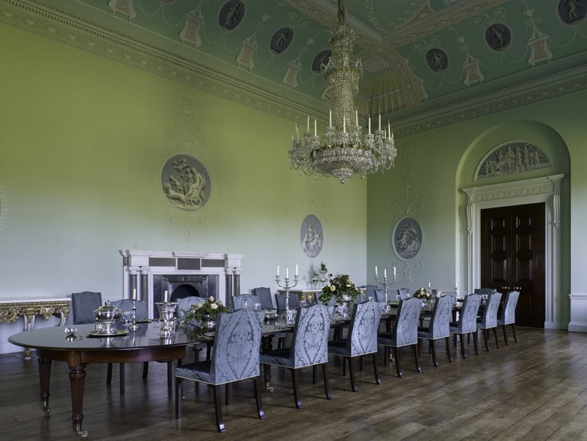 Elegant dining room with long table, ornate chandelier, high ceiling, decorative moldings, and classical wall art.