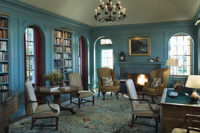 Cozy library with blue walls, chandelier, fireplace, bookshelves, and comfortable seating with patterned rug.