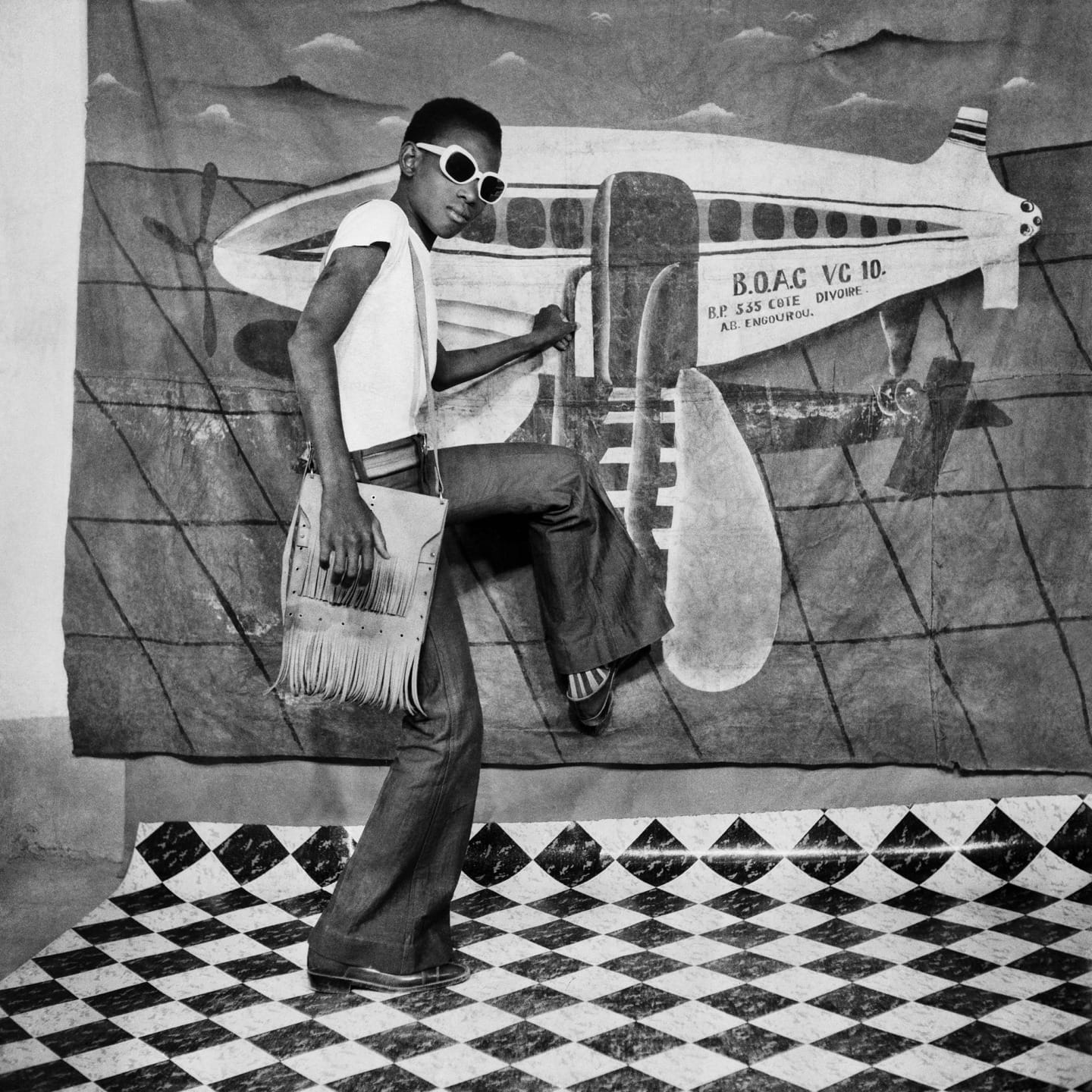 Person in sunglasses and flared jeans poses in front of airplane backdrop on checkered floor holding a fringed bag.