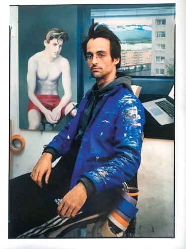 Artist in a blue jacket seated in a studio with a painting and laptop in the background.