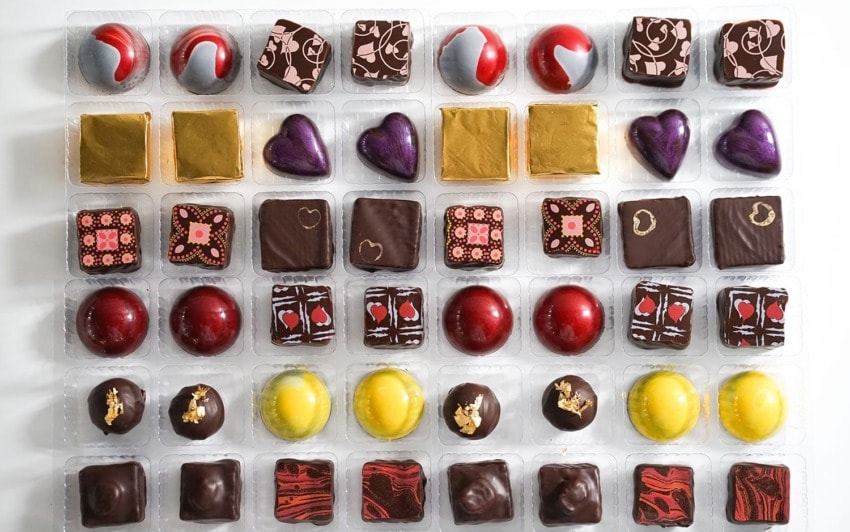 Assorted gourmet chocolates with various shapes and decorative toppings arranged in a grid pattern on a white background.