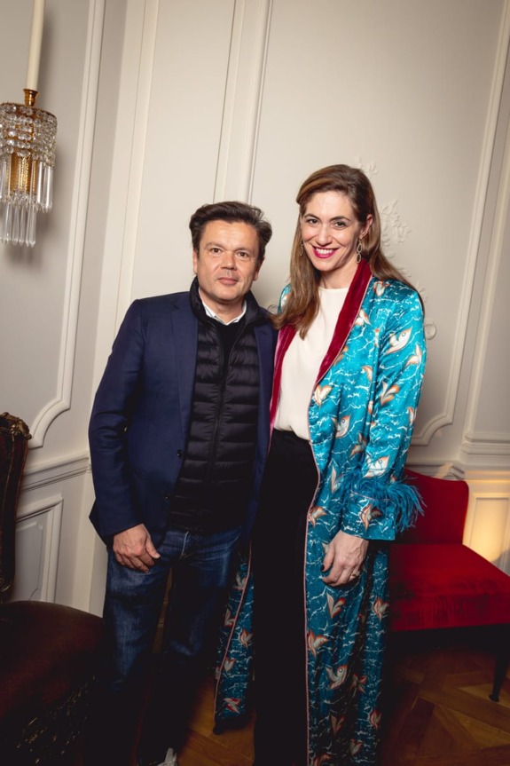 Two people posing in a stylish, elegantly decorated room with a chandelier and ornate wall details.