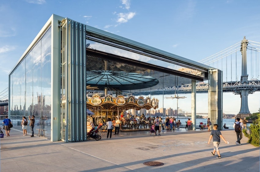 Modern glass building housing a carousel with a bridge and city skyline in the background, people walking nearby.