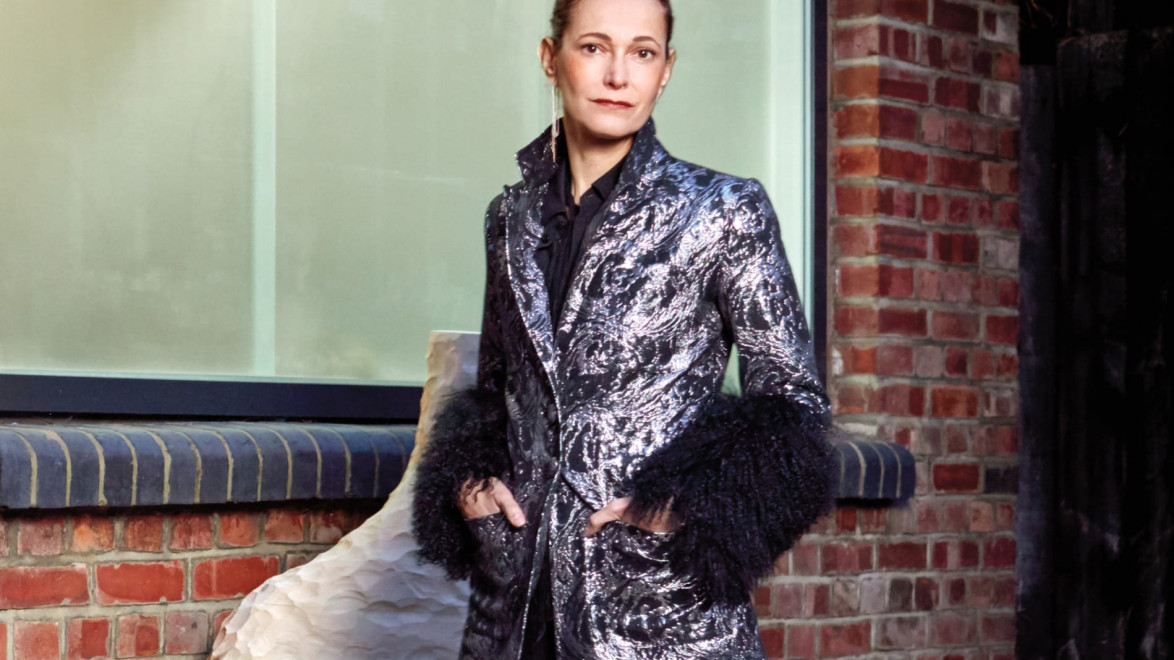 Person wearing a patterned coat stands outside a brick building near a large window.