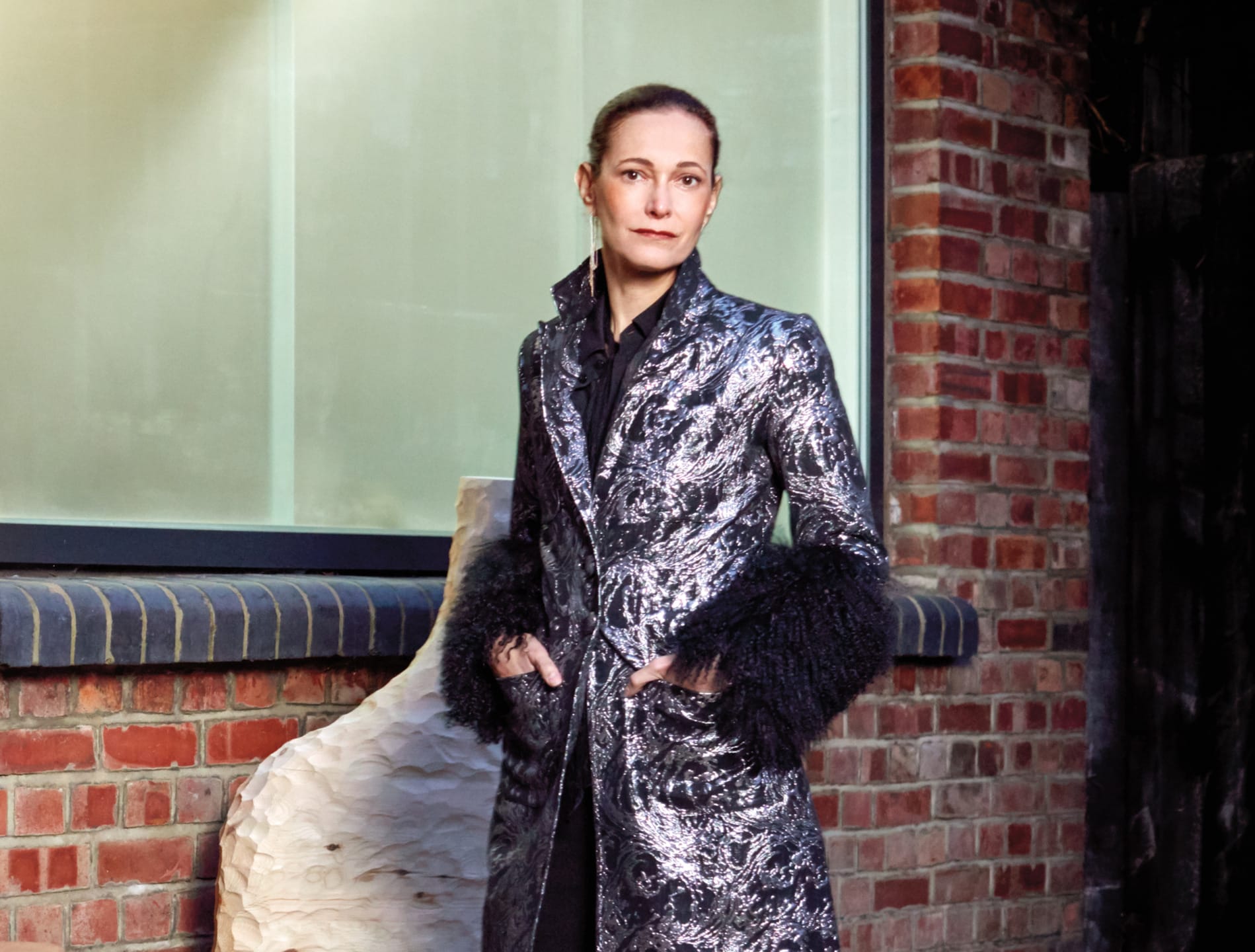 Person wearing a patterned coat stands outside a brick building near a large window.