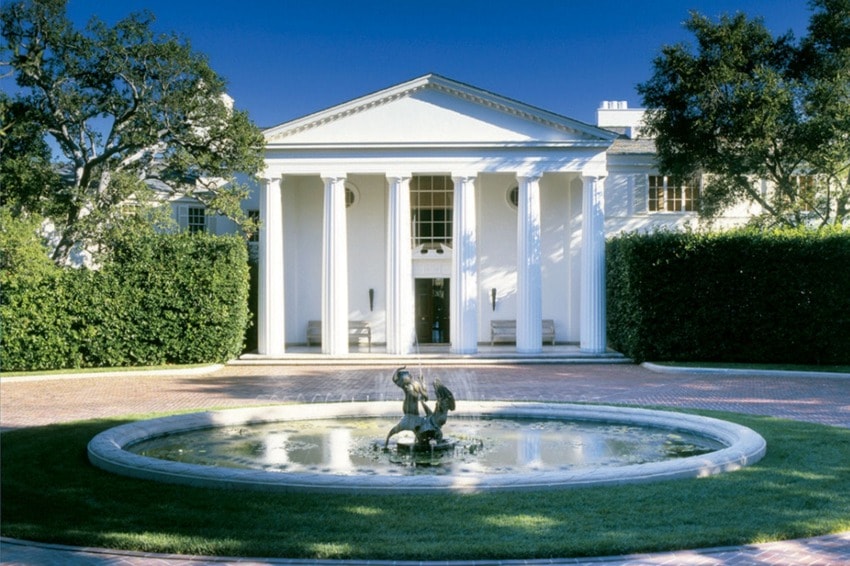 Exterior view of a classic white building with columns and a central fountain surrounded by greenery and trees under a clear sky.