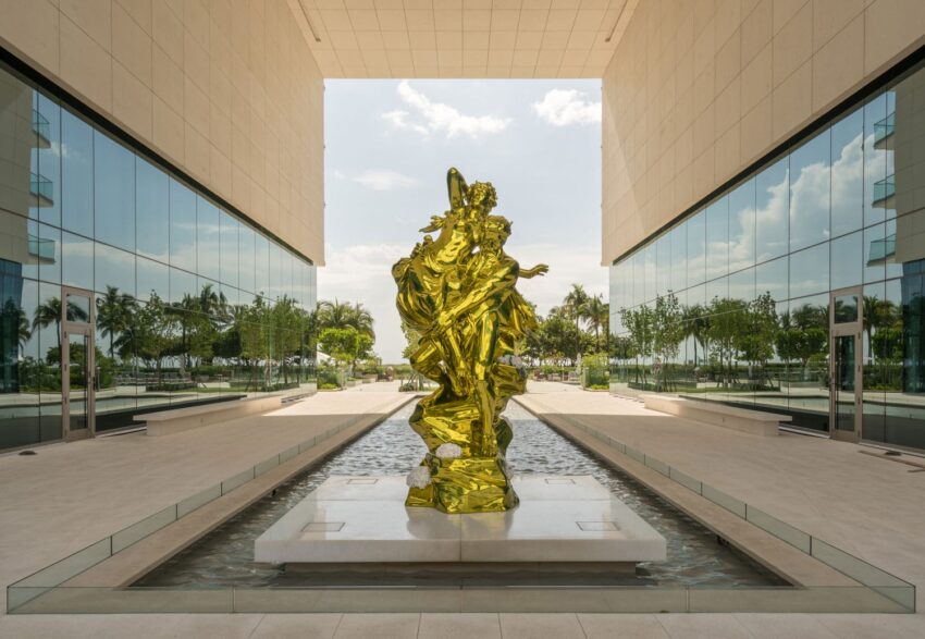 Golden sculpture in modern courtyard with reflective pool and glass building, surrounded by trees and cloudy sky.