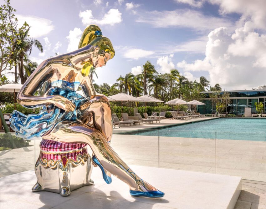 Colorful reflective sculpture of a seated ballerina near a luxurious poolside with palm trees and lounge chairs in the background.