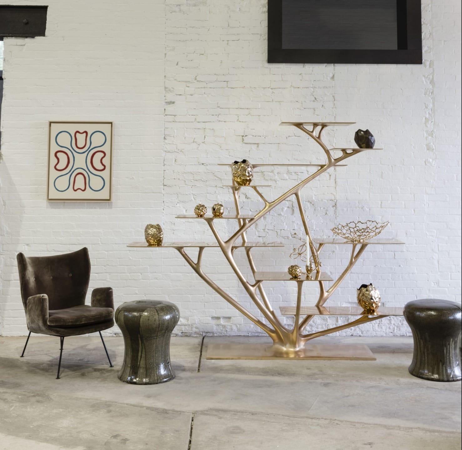 Modern art installation with a metallic tree structure holding decorative objects, flanked by a chair and stools against a white wall.
