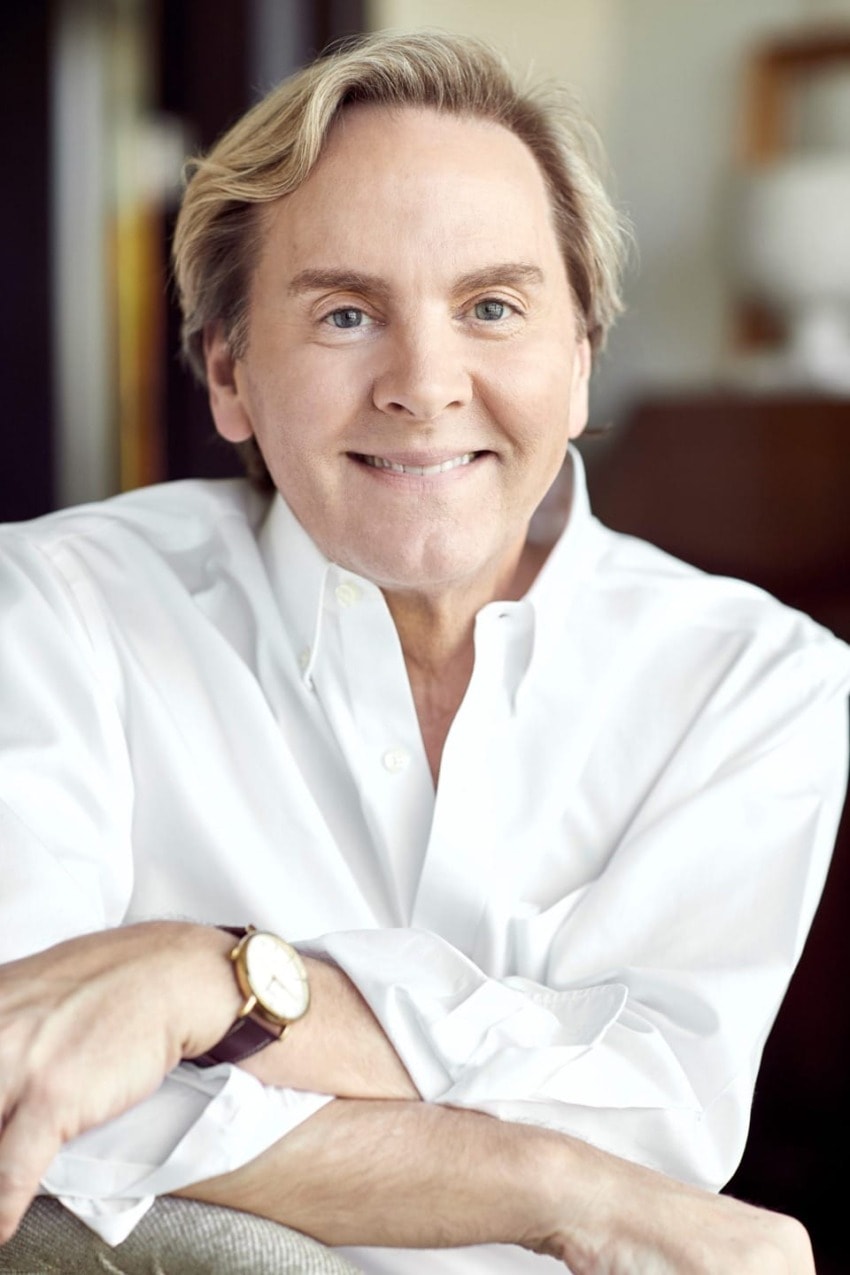 Man smiling in a white shirt, sitting with a watch on his wrist, in a relaxed indoor setting.