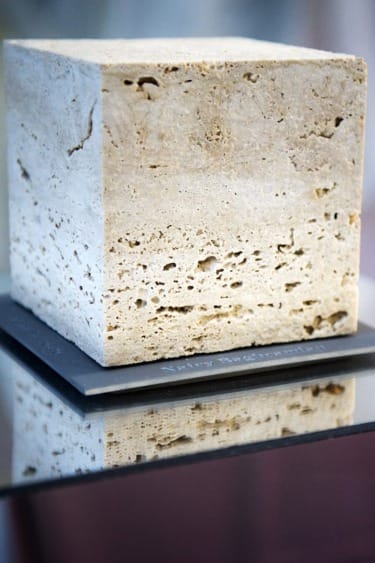 A large cube of travertine stone displayed on a reflective surface showing its textured surface and natural patterns.